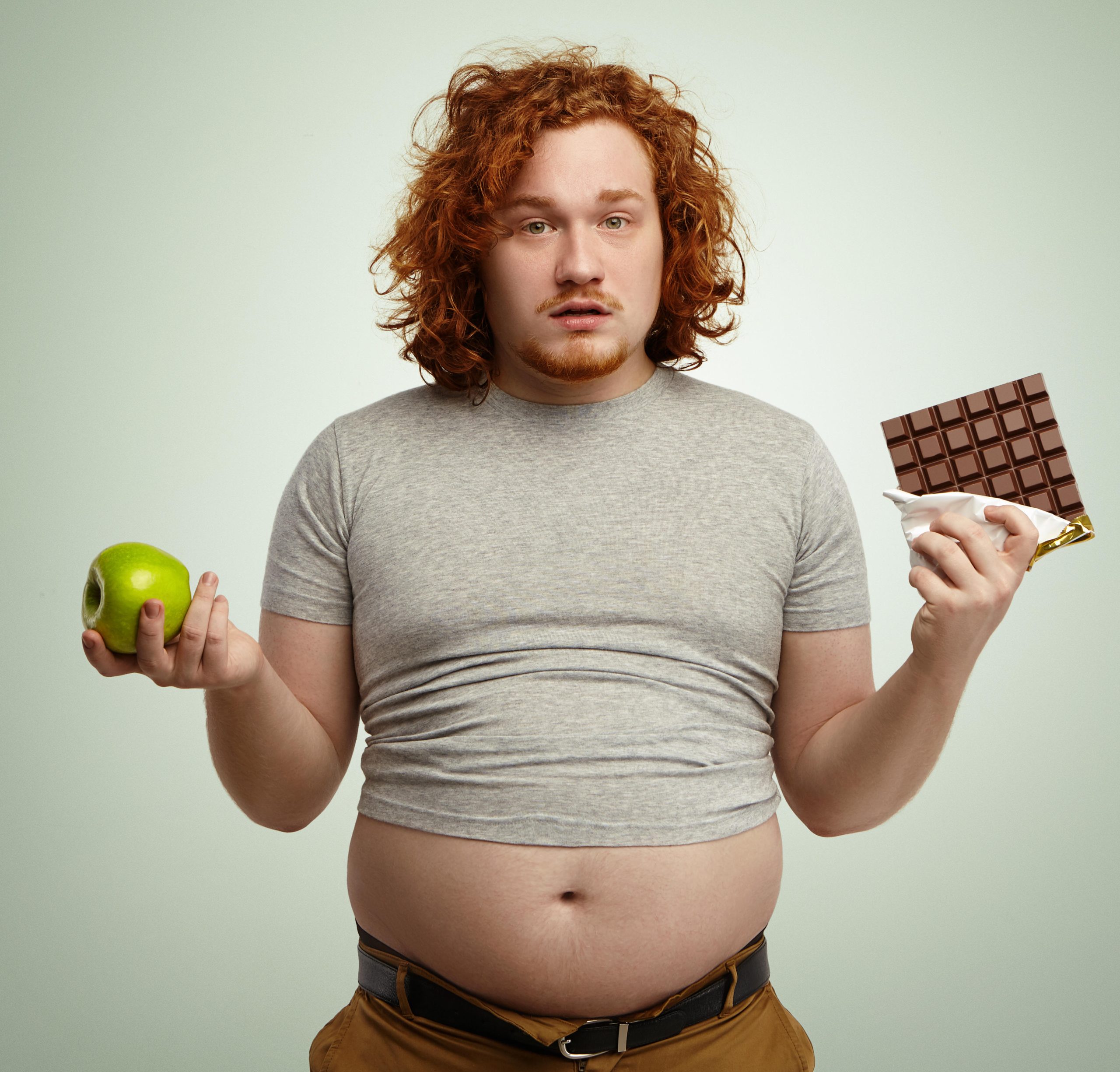 overweight man with chocolate in one hand and an apple in the other with red hair looking like he doesn't understand the reasons he is not losing weight.