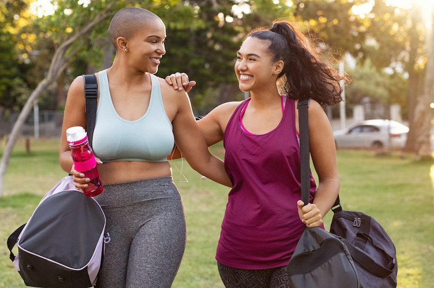 An overweight black woman in a blue tanktop and a hispanic woman in an purple shit walking with arms around each other happy for healthy living