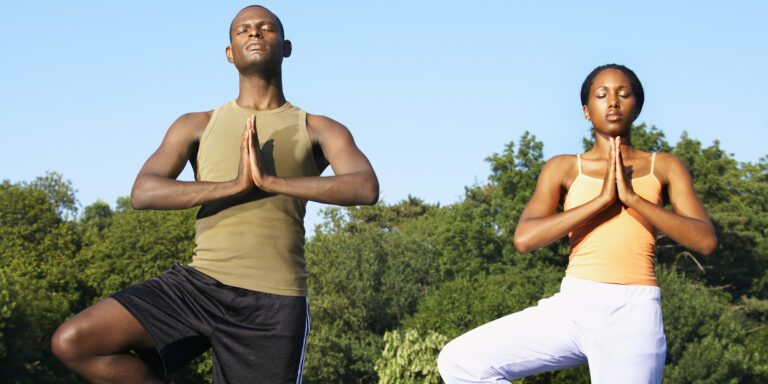 An black man and woman in a green shirt and yellow tank top standing on one leg and arms in a praying position praying for improved health and wellness while doing yoga