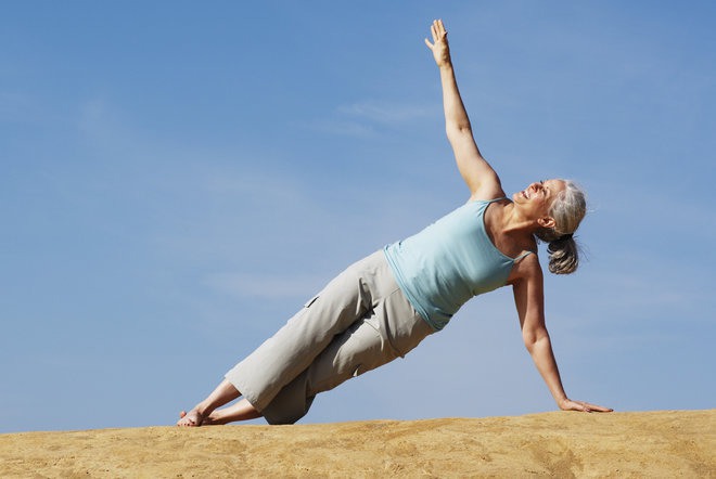 Elderly lady in a blue tank top and grey pants on one arm leaning doing yoga to improve health and wellness